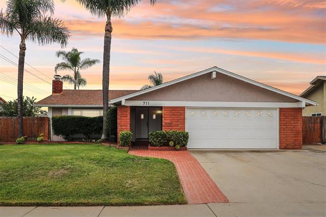 Single-story home with a lit front window, a lawn and palm trees at dusk